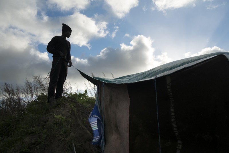 Dougar* Baha, migrant soudanais, pose la bâche qui servira de toit à la nouvelle habitation qu'il vient de construire. 
Cet espace servira de cuisine à sa communauté.