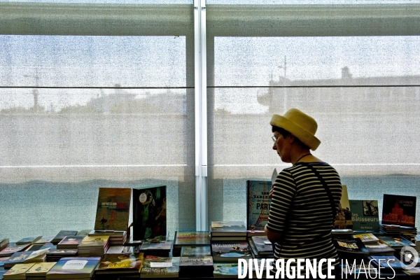Illustration Aout 2016.Dans la boutique du musee d art moderne une femme regarde les livres proposes a la vente.En silhouette derriere le store, un paquebot a quai